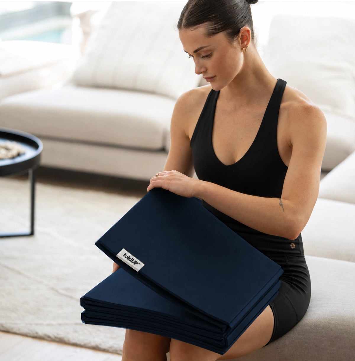 Woman seated on a sofa folding a navy Movement Mat into a compact stack on her lap.