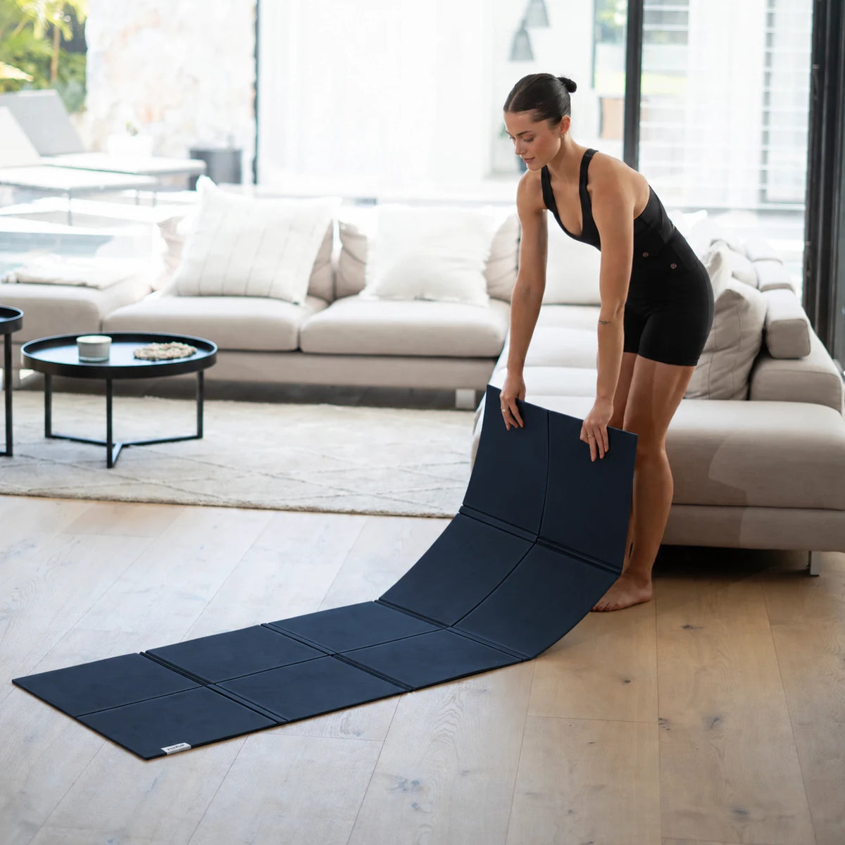 Woman unfolding a navy Movement Mat on a timber floor beside a sofa.