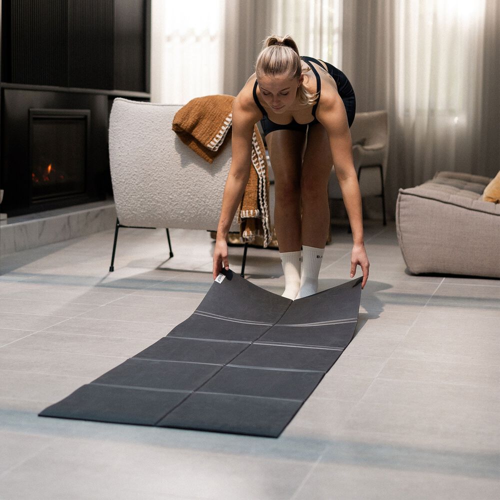 Woman laying a black Movement Mat flat on the floor in front of a living room armchair.