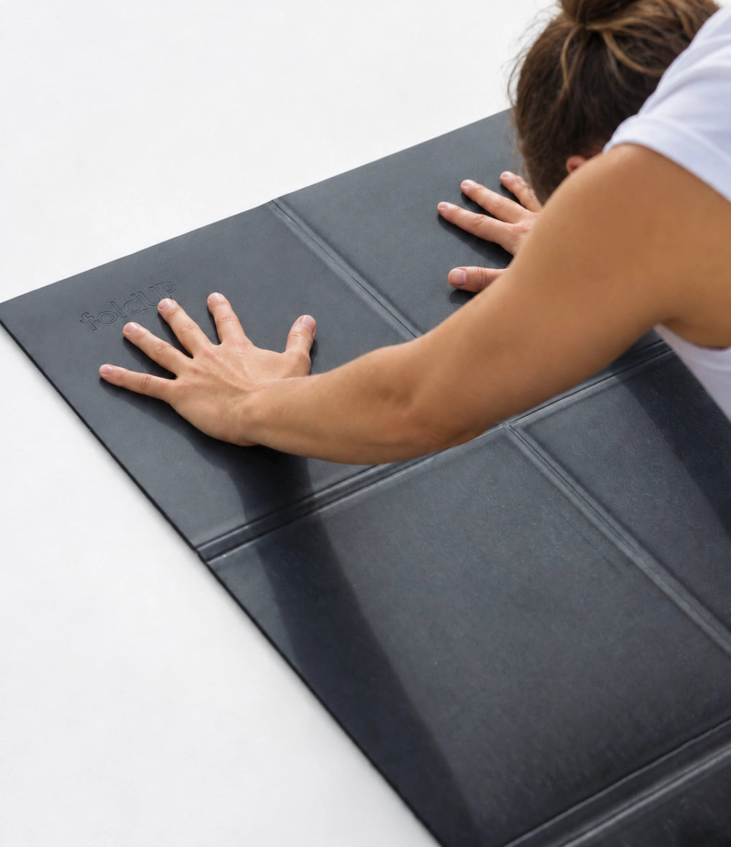 A person in a white tank top pressing down on a foldup black movement mat, multi-panel yoga mat on a clean white surface.