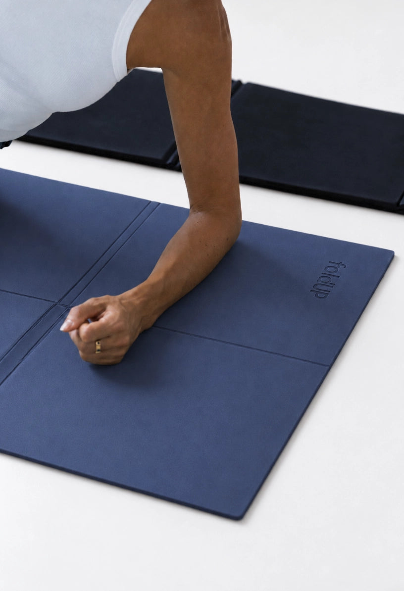 A person in a white sleeveless top performs a plank on a midnight blue foldup movement mat, with a folded navy mat and a black mat in the background on a white floor.