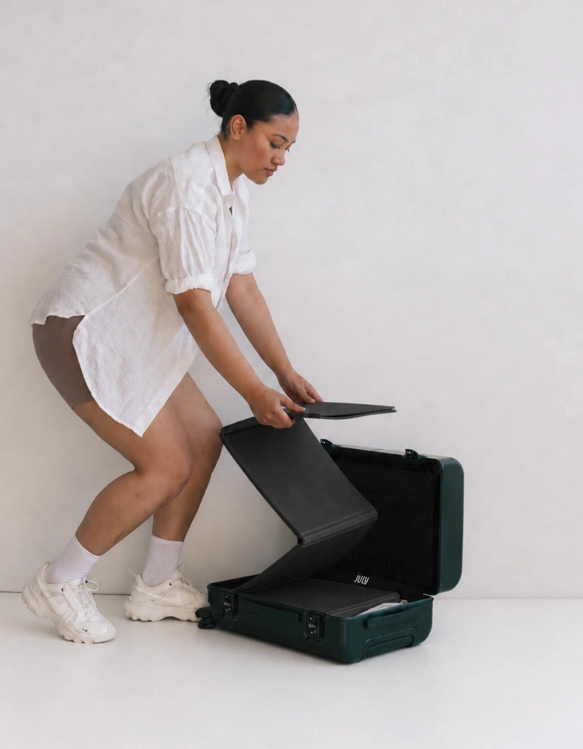 A woman in a white shirt and brown shorts is crouching down to place a black foldup movement mat into an open dark green hard-shell laptop suitcase on a white studio floor.