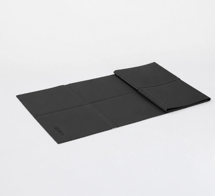 A black foldable yoga mat shown partially unfolded on a clean white studio background, with a matching black sleeve or pouch placed on top.
