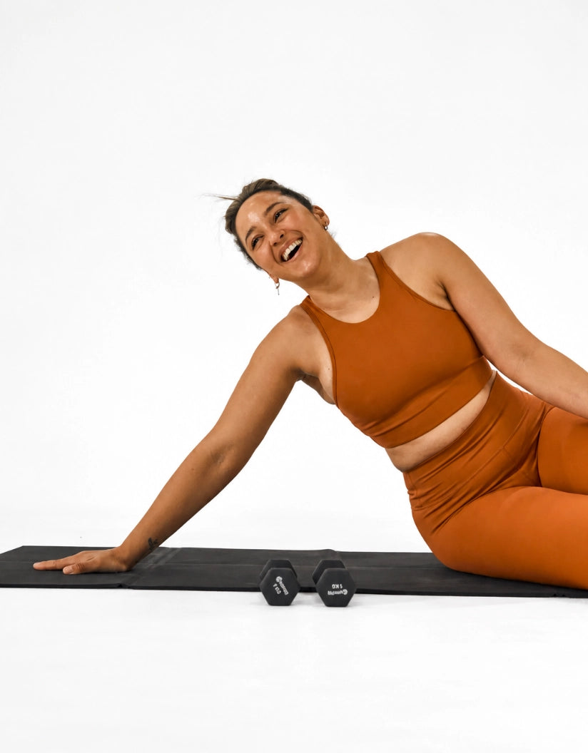 A woman in a matching burnt orange activewear set lies on a black yoga mat branded 'foldup' on a white studio floor, holding a small black hex dumbbell. She is smiling and looking to the side.