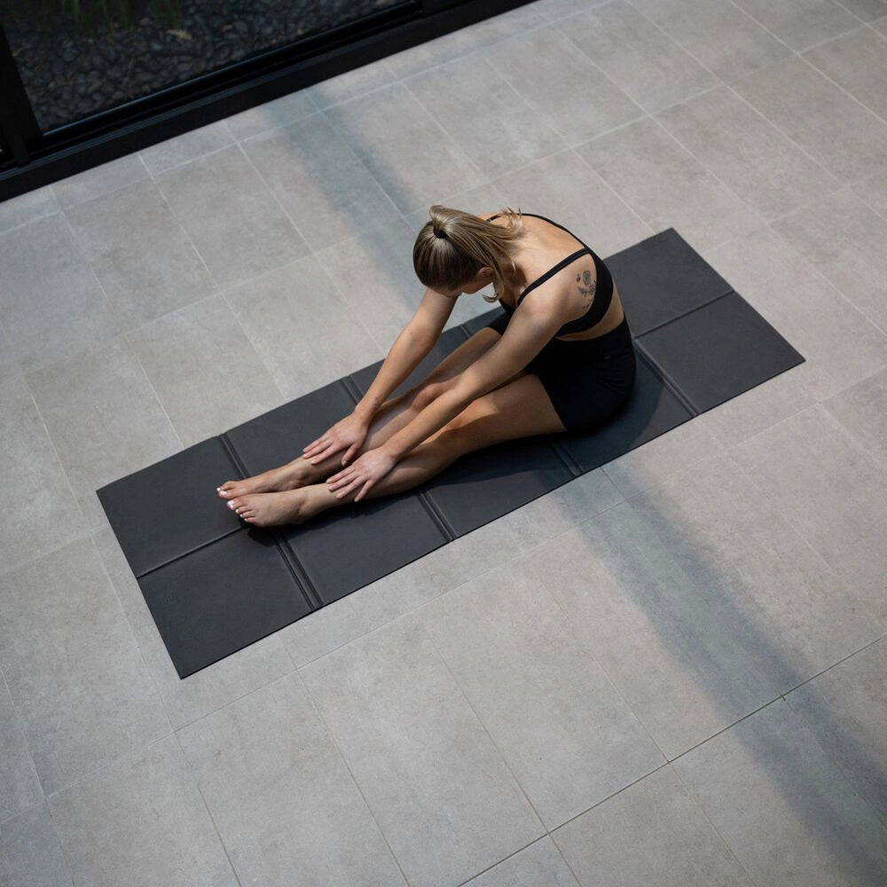 Overhead view of a woman reaching towards her feet in a seated stretch on a black Movement Mat.