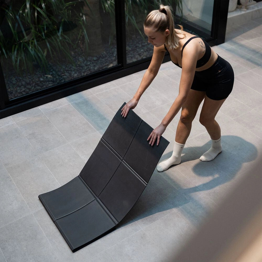 Woman unfolding a black Movement Mat on a tiled floor beside a large window.